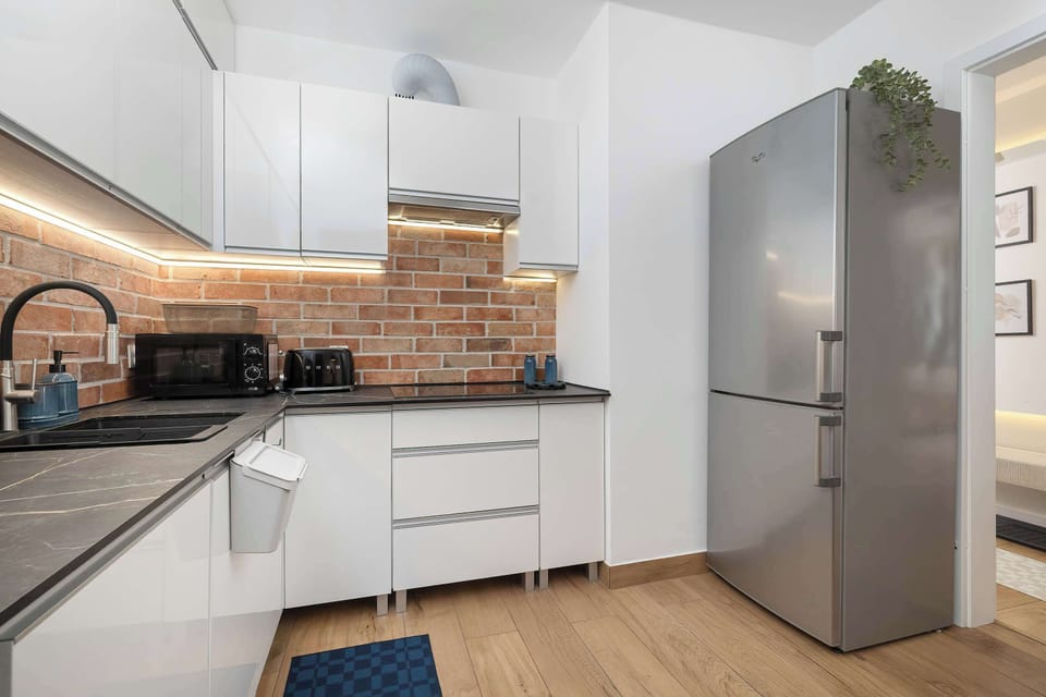 A narrow kitchen layout showcases a stainless steel refrigerator standing next to white cabinetry and a brick backsplash. Clean countertops and under-cabinet LED lighting enhance the modern feel.