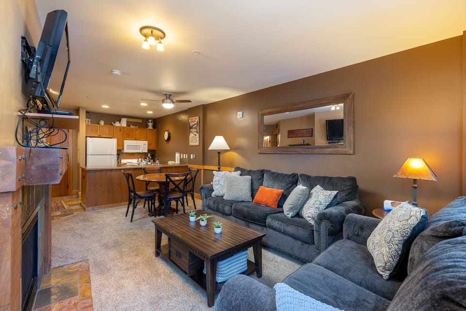 A cozy living room features a large gray sectional sofa with multiple pillows, a wooden coffee table, a wall-mounted TV, and a dining area with a kitchen in the background. Ambient lighting illuminates the space.