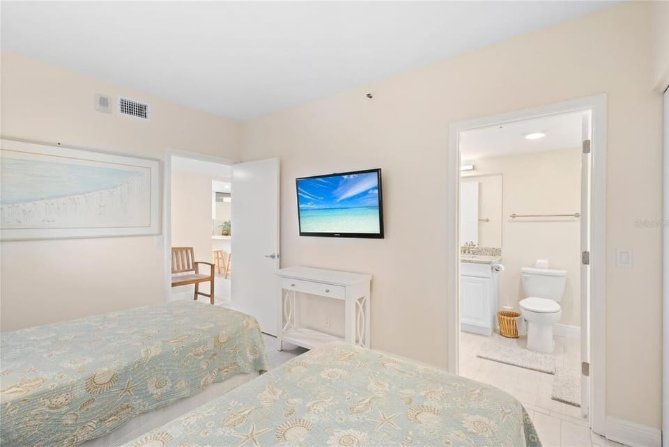 Twin beds with seashell-patterned quilts face a wall-mounted TV, with a bathroom visible through an open door.