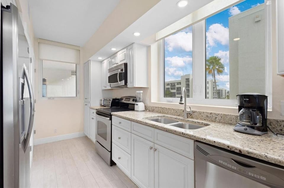 Galley kitchen with granite countertops, white cabinetry, stainless steel appliances, and a view through large windows.
