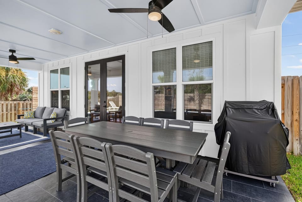 Covered patio with dark wood dining table, eight chairs, gray cushioned sofa, ceiling fans, and a black grill.