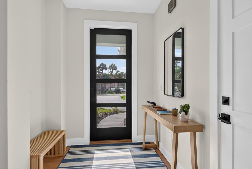 Entryway with black-framed glass door, natural wood console table, slatted bench, and striped blue rug.