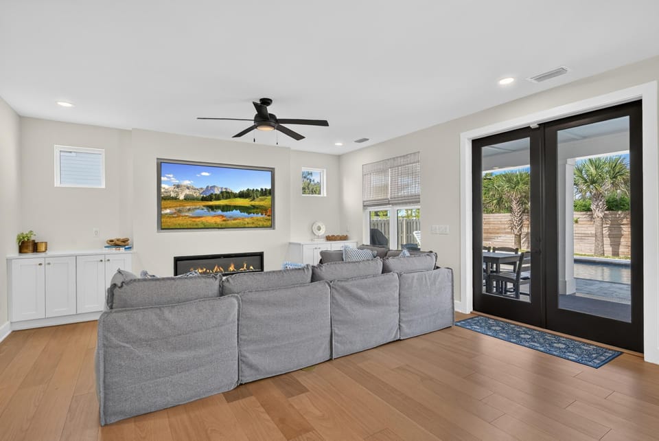 Living room with light wood floors, a gray sectional facing a modern gas fireplace, and black-framed glass doors to the pool.