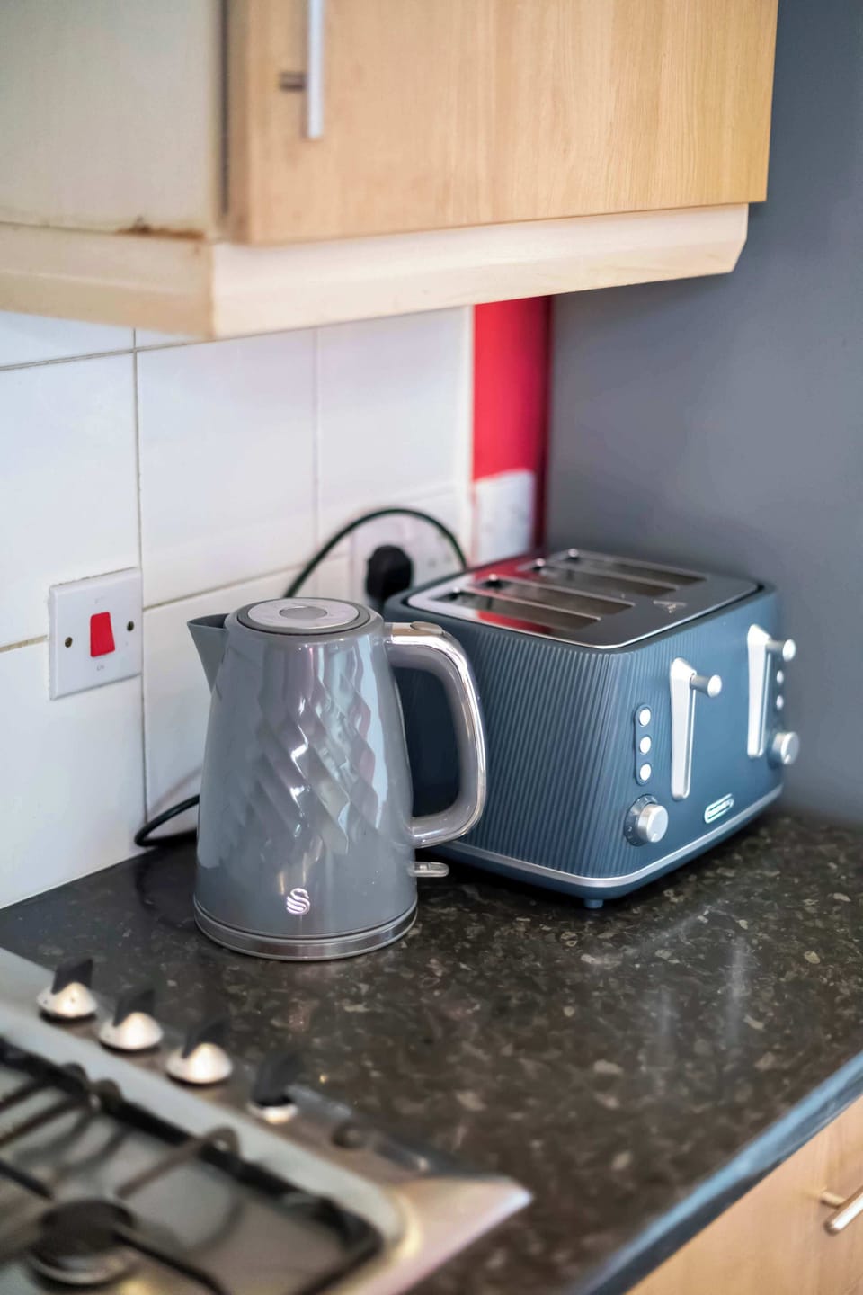 This kitchen countertop setup features a gray electric kettle and a matching blue toaster next to a gas stovetop.