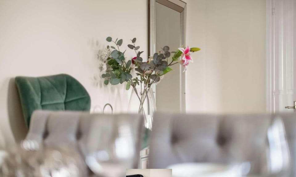 This dining and seating area includes comfortable gray tufted chairs, an accent green chair, and a clear flower vase placed in front of a tall mirror.