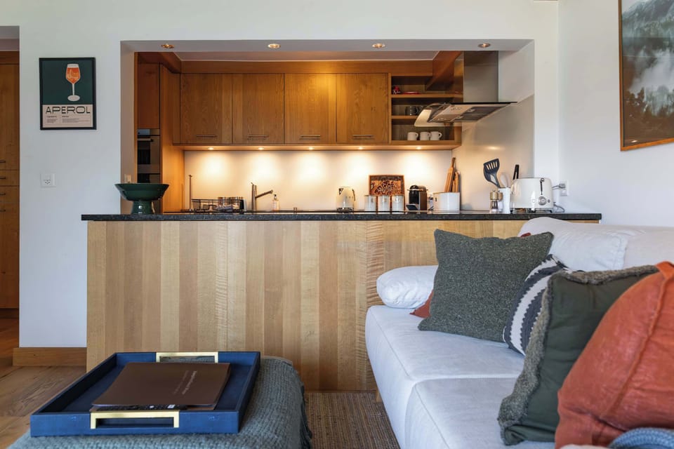 Prepare meals easily in this fully equipped open kitchen, featuring warm wooden cabinetry, modern appliances, under-cabinet lighting, and a breakfast counter directly facing the cozy sofa lounge.