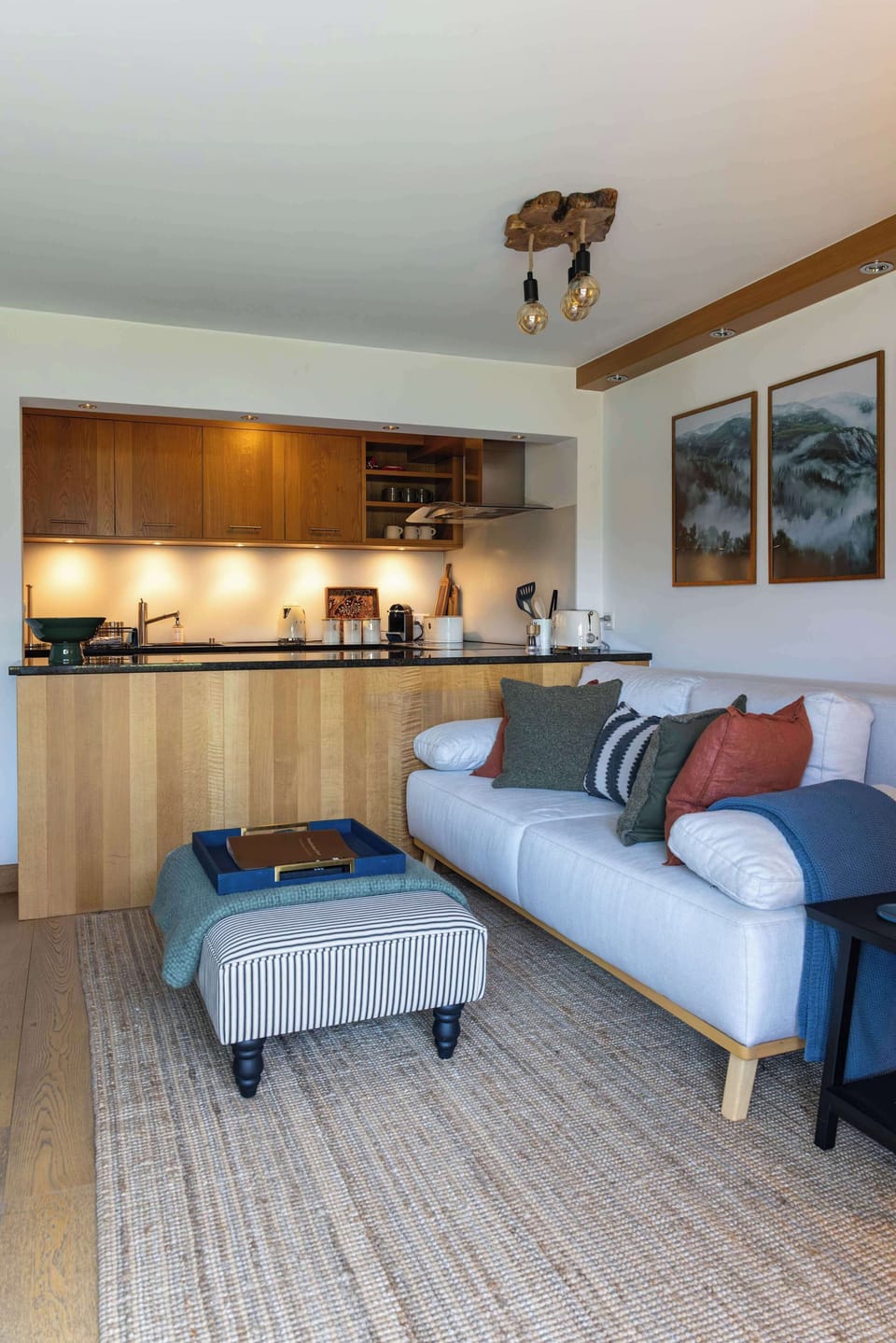 Unwind in this stylish lounge space, featuring a comfortable white sofa with colorful throw pillows, a striped ottoman over a textured rug, scenic mountain wall art, and an open layout directly connected to the modern kitchen.