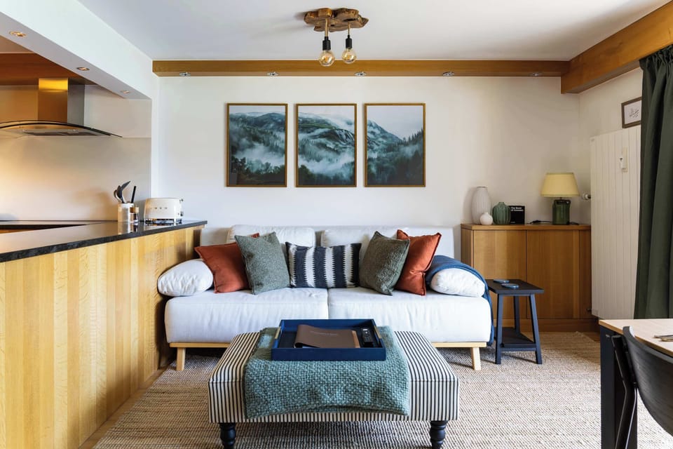 Relax in this cozy living room nook, featuring a plush white sofa, a striped ottoman coffee table, mountain art triptychs, and an adjacent kitchen counter.
