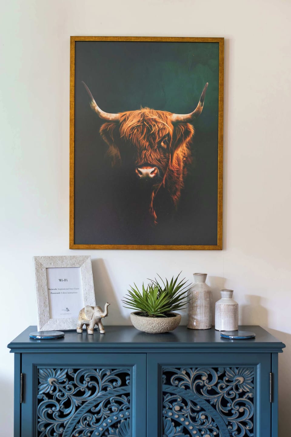 Admires the charming decor setup, featuring a framed portrait of a Highland cow mounted above an ornate blue console table decorated with a Wi-Fi frame, a elephant figurine, and potted greenery.