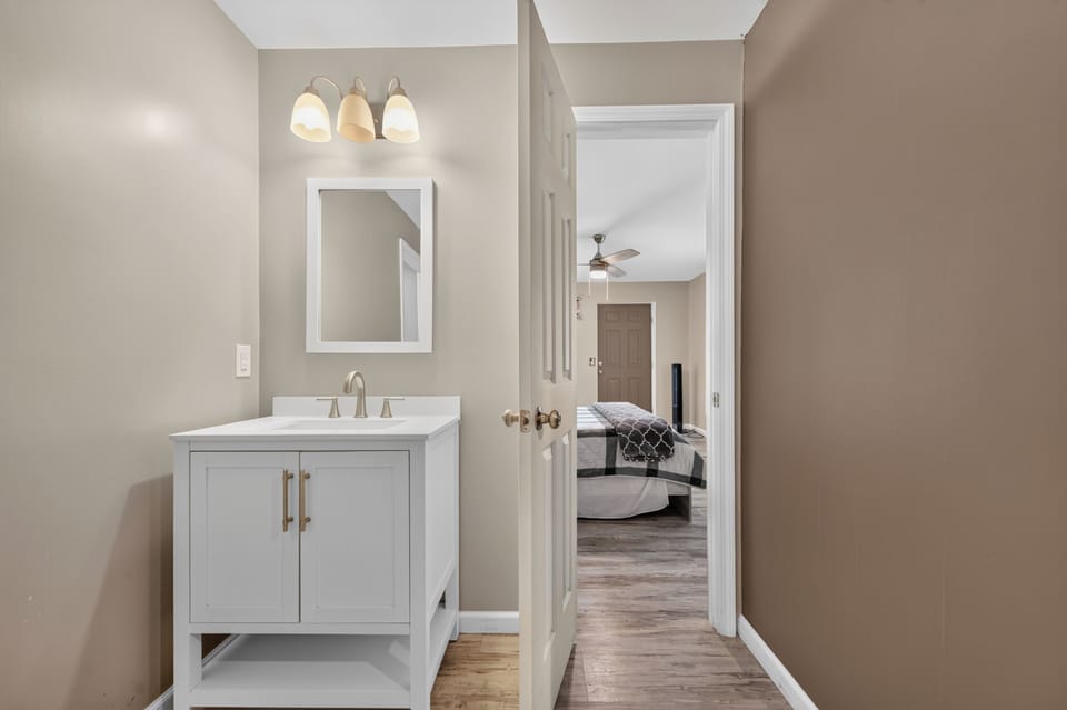 A bathroom with a white vanity and mirror, with an open door looking into a bedroom.