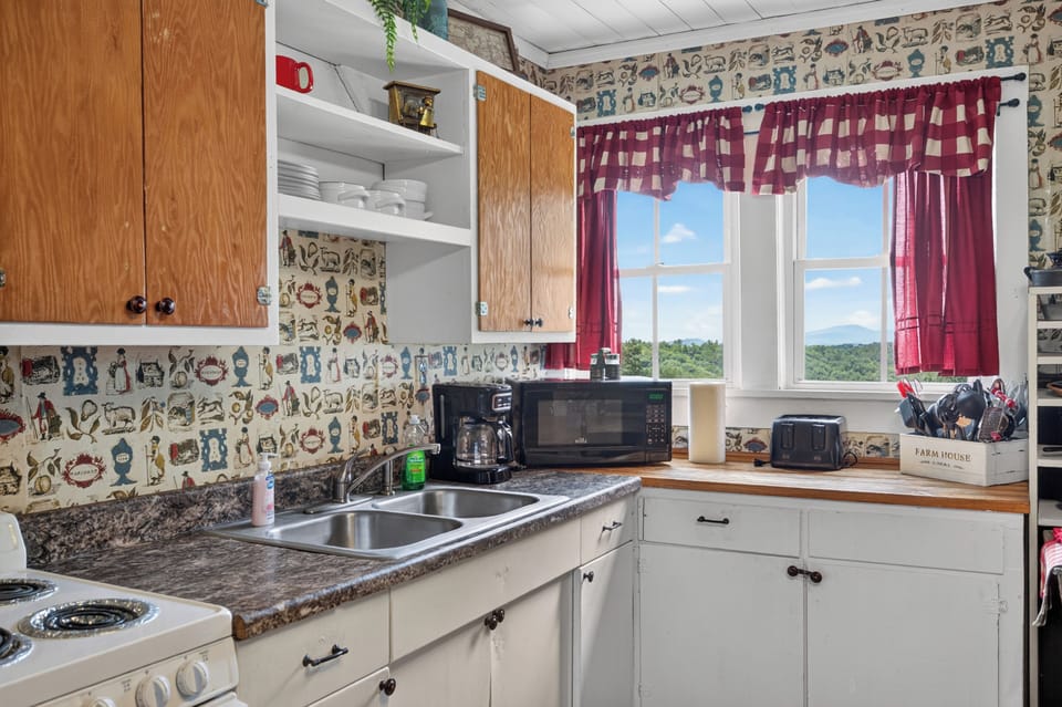 A rustic kitchen counter with wood-front cabinets, vintage wallpaper, and a window overlooking a scenic view.
