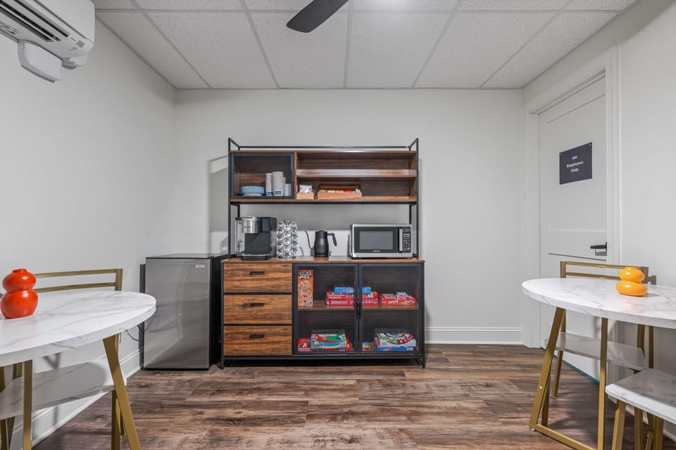 Shared refreshment station with a microwave, mini-fridge, coffee maker, glassware, and board games.