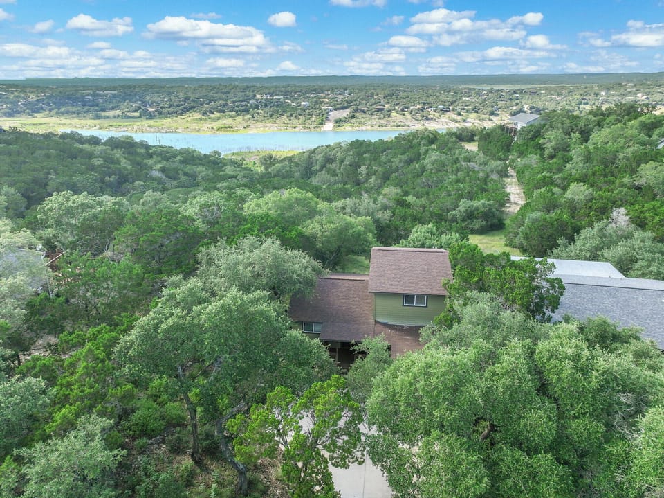 Aerial view of the property nestled among lush trees with a serene lake backdrop, offering a peaceful natural setting for a relaxing Hill Country retreat.