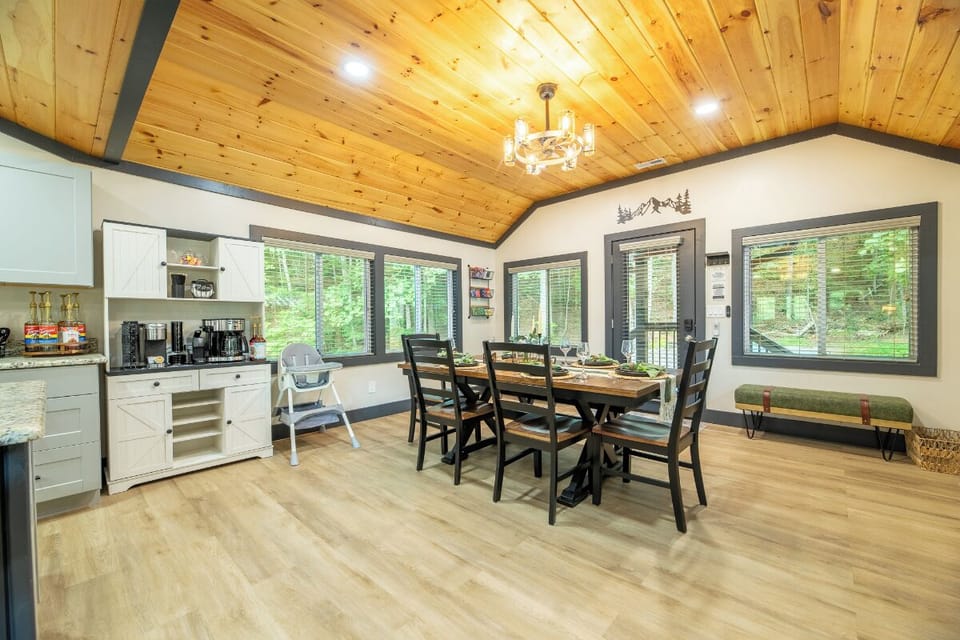 Gather around the rustic dining table beneath warm wood-paneled ceilings, where natural light streams through windows revealing peaceful forest views beyond your cozy retreat.