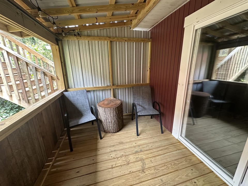 Covered porch with wood plank flooring, metal siding, two gray mesh chairs, and a tree stump side table.