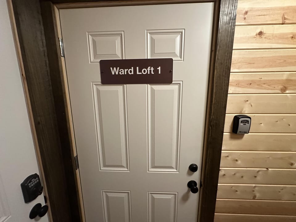 White paneled door with a dark brown sign reading "Ward Loft 1," framed by natural wood walls and a keypad lock.