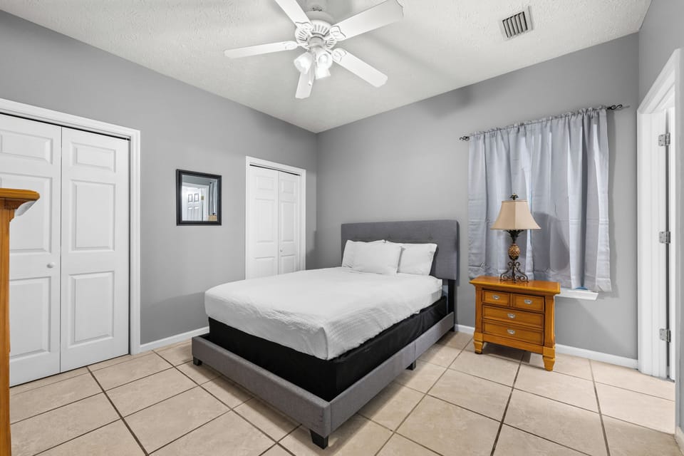 Your peaceful bedroom features a plush queen bed dressed in crisp white linens, with a ceiling fan overhead to keep you cool. Natural light filters through the curtains, creating a restful space perfect for unwinding.