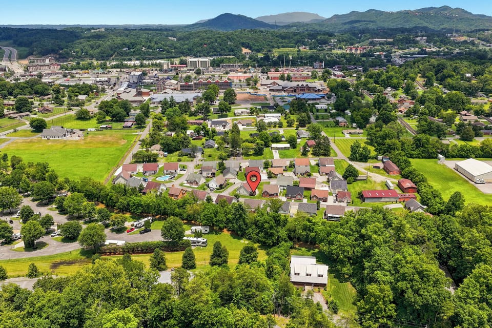 Aerial view of a peaceful residential neighborhood nestled among green fields and trees, with mountain ranges visible in the distance and a charming small town center nearby.