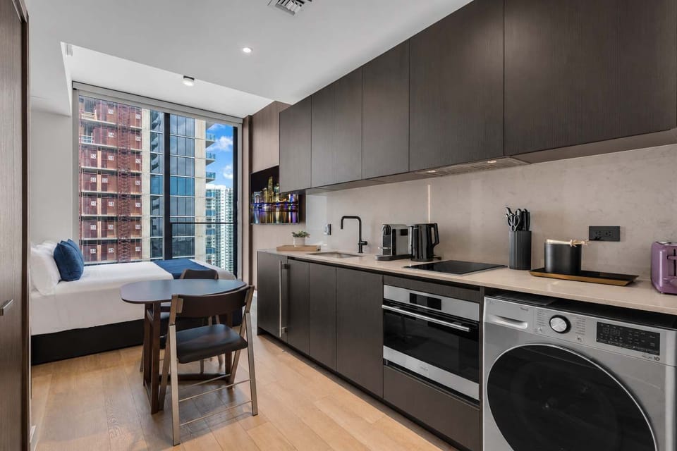 Open kitchen and dining nook with full appliances, coffee maker, and washer/dryer. A bistro table sits by the window, with a built-in daybed and city views just beyond.