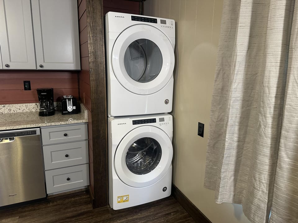 Stacked white Amana washer and dryer sit next to gray kitchen cabinets, granite countertops, and a beige curtain.