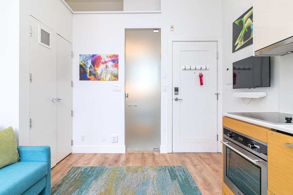 Entryway with frosted glass door, white walls, colorful abstract art, and adjacent kitchen cabinetry in light wood finish.