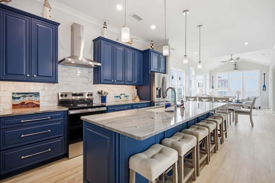 Stunning gourmet kitchen with navy cabinetry, oversized quartz island seating for six, stainless steel appliances, marble backsplash, pendant lighting, and an open layout flowing to the dining area and ocean view great room.