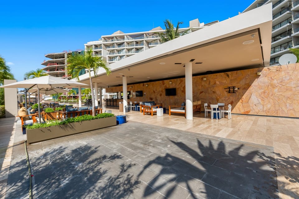 Enjoy this expansive covered outdoor pavilion, featuring comfortable seating areas, flat-screen TVs, a stone accent wall, a chic bar setup, and sunny poolside terraces.