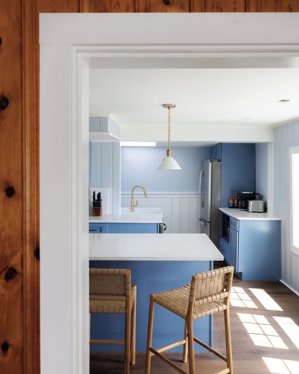 Blue-gray island and rattan stools make the kitchen a natural gathering spot even before the meal — someone always ends up perched here all evening.