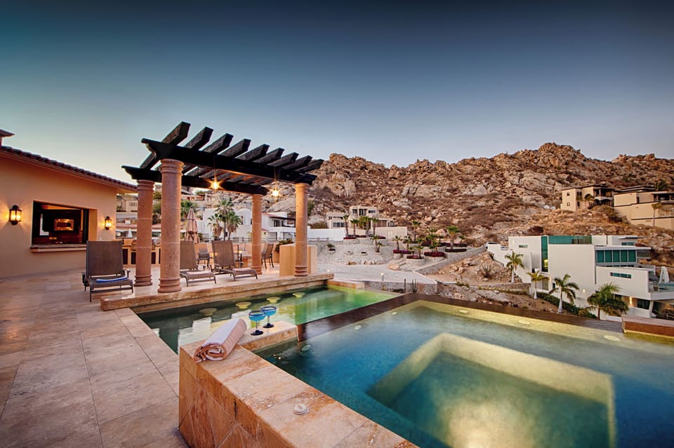 A dramatic hillside property with a multi-level pool and pergola-covered terrace, set against rugged desert mountains at dusk.