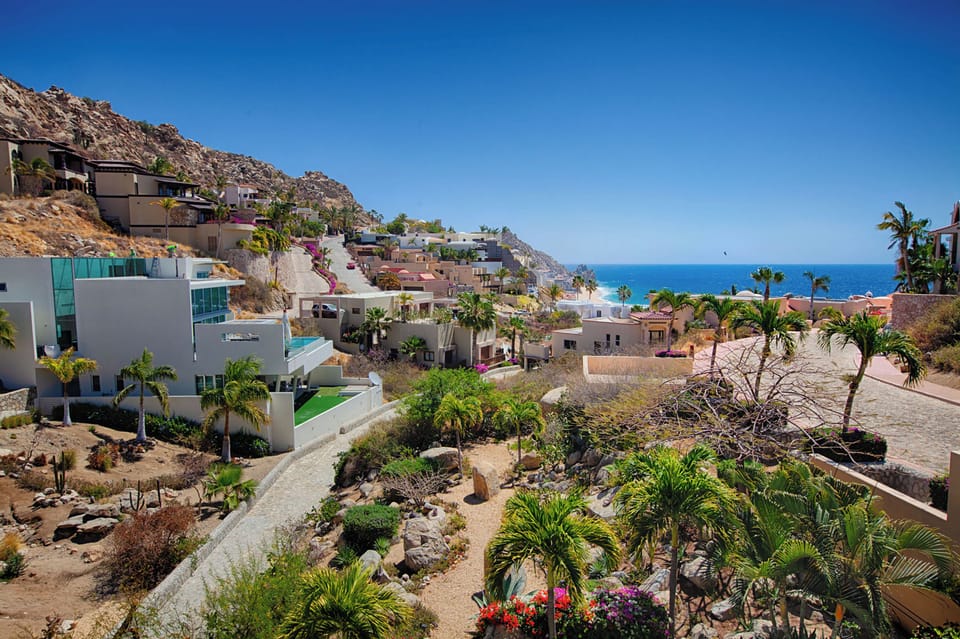 Coastal hillside community featuring terraced homes overlooking the ocean, with palm trees dotting the rocky landscape and a winding pathway through landscaped gardens. The property area showcases colorful architecture against dramatic blue skies.