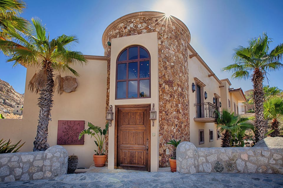 Mediterranean-style villa with distinctive stone turret entrance, arched wooden door, and palm-lined courtyard in a hillside setting.