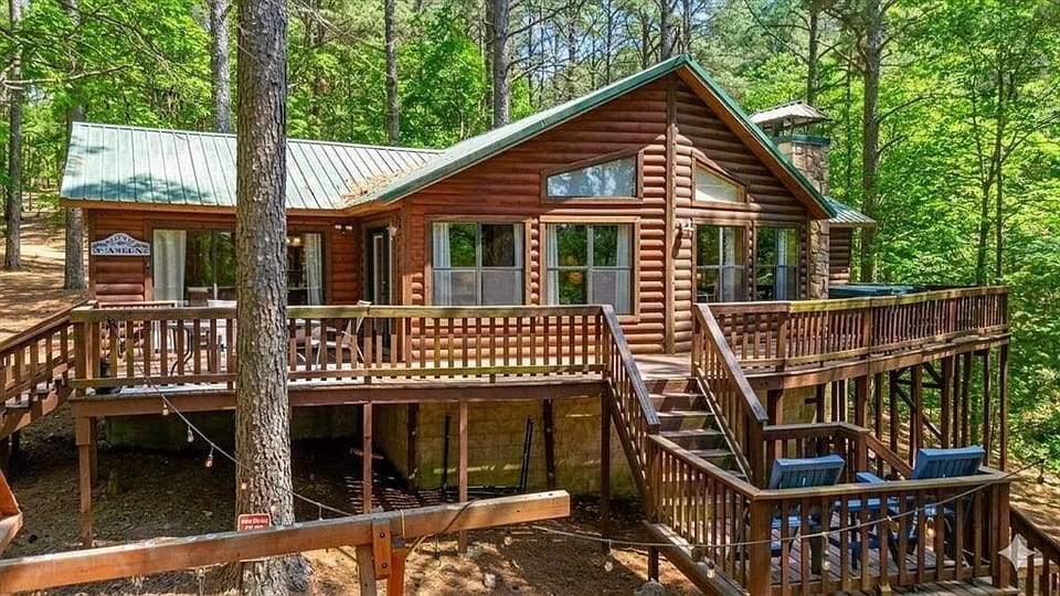 A charming log cabin nestled in a lush forest setting, featuring a spacious wraparound deck perfect for outdoor relaxation and forest views.