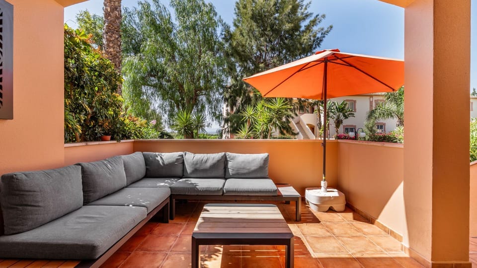Unwind in ultimate comfort on this expansive patio, lounging on the stylish outdoor sofa under the shade of a vibrant orange parasol, perfect for enjoying al fresco meals or simply soaking up the sun in privacy.
