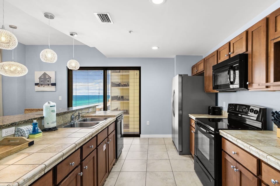 Your fully equipped kitchen has everything you need—stainless steel appliances, rich wood cabinetry, and stunning ocean views through the glass door, blending convenience with coastal beauty for your perfect getaway.
