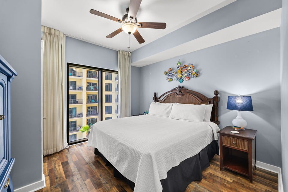 Unwind in your serene bedroom featuring a plush bed with crisp linens, warm wood furnishings, soothing blue walls, and a ceiling fan to keep you comfortable throughout your stay.