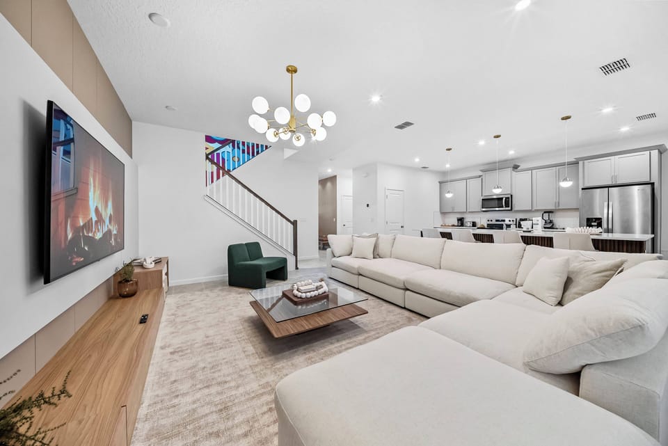 Enjoy the bright and airy living room with a spacious sectional sofa and an elegant chandelier. The open kitchen, featuring sleek cabinetry and stainless steel appliances, invites you to relax and feel at home.