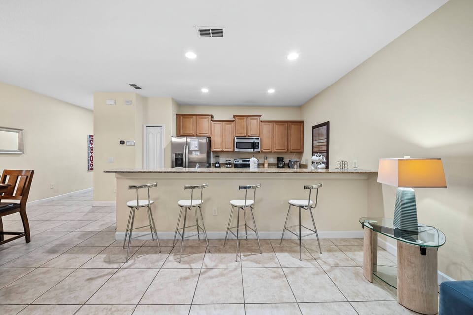 This bright kitchen features sleek wood cabinetry and a spacious bar with stylish metal stools, perfect for casual meals. The tiled flooring and warm beige walls create a welcoming atmosphere for your culinary adventures.