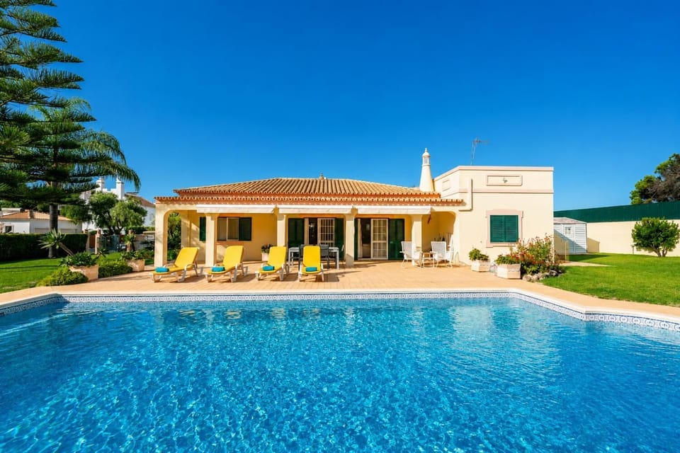 Beautiful single-story villa with a large swimming pool, tiled patio, and yellow lounge chairs.