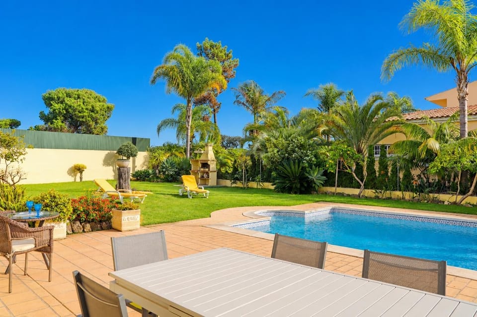 A poolside view featuring a spacious patio with dining furniture in the foreground, looking across a swimming pool toward a lawn with palm trees and a brick barbecue grill.