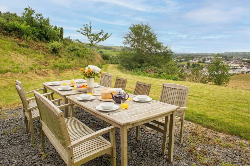 Outdoor wooden dining table set for six on gravel, with plates and food, overlooking a grassy hillside