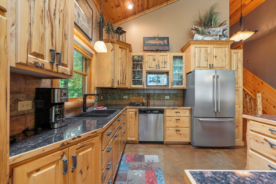 Your fully-equipped kitchen blends rustic charm with modern convenience, featuring custom wood cabinetry, gleaming granite counters, and stainless appliances beneath warm wood-paneled ceilings and natural light.