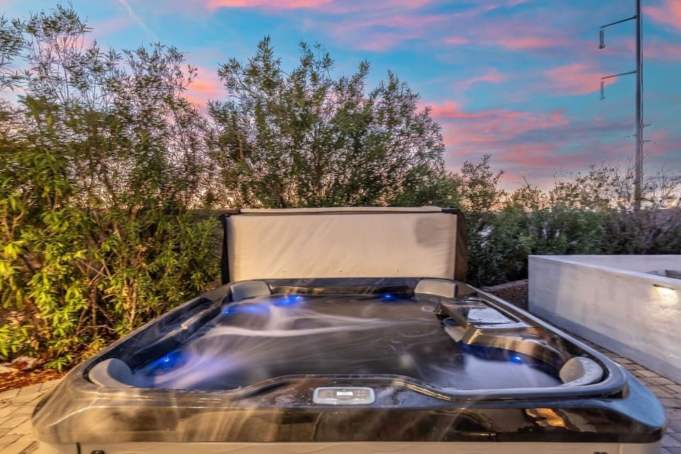 A steaming hot tub glows under a pastel desert sunset, tucked beside lush greenery for total privacy.
