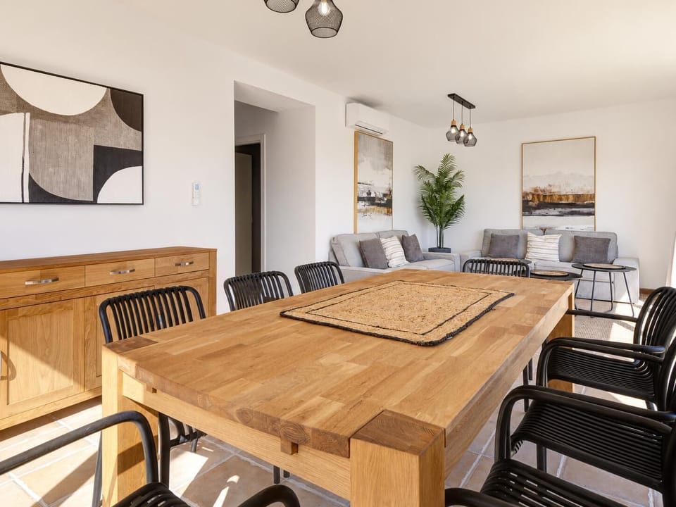 Spacious, open-plan dining and living area featuring a large wooden dining table with stylish black chairs, modern grey sofas, contemporary artwork, and a warm wooden credenza.