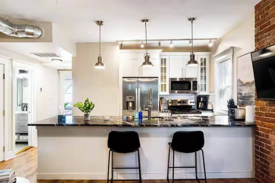Enjoy the perfect blend of comfort and style in this bright, open-concept living area featuring a modern kitchen, cozy seating, and charming exposed brick accents.