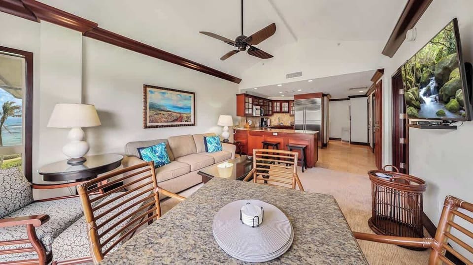 Relax in your open-concept living area where rich wood accents and tropical decor create a warm island vibe. Unwind on the plush sofa, catch ocean views through the doors, and enjoy the fully equipped kitchen with modern appliances.