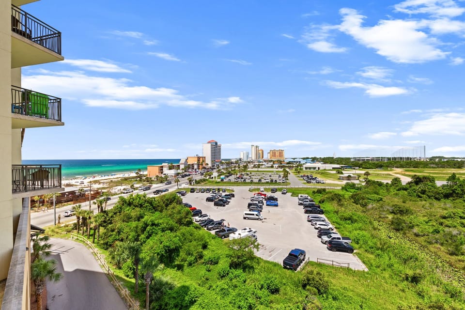 A coastal property with balconies overlooking the turquoise Gulf waters, beachside buildings, and convenient parking below. The surrounding area features palm-lined paths and nearby city towers.