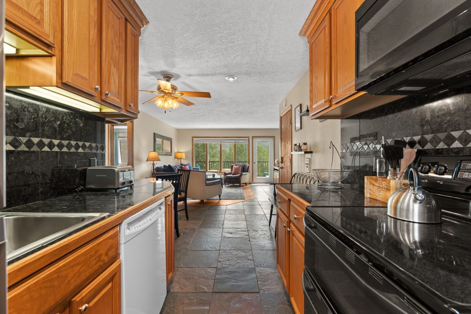 Your spacious kitchen features gleaming granite counters, warm wood cabinetry, and modern appliances—perfect for preparing meals while enjoying views of the cozy living area and sunlit deck beyond.