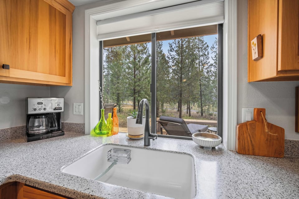 Picture yourself preparing meals in this bright kitchen, where sunlight streams through the window and views of towering pines bring the outdoors in. Natural wood cabinetry and a sleek workspace create a cozy, functional setting for your stay.