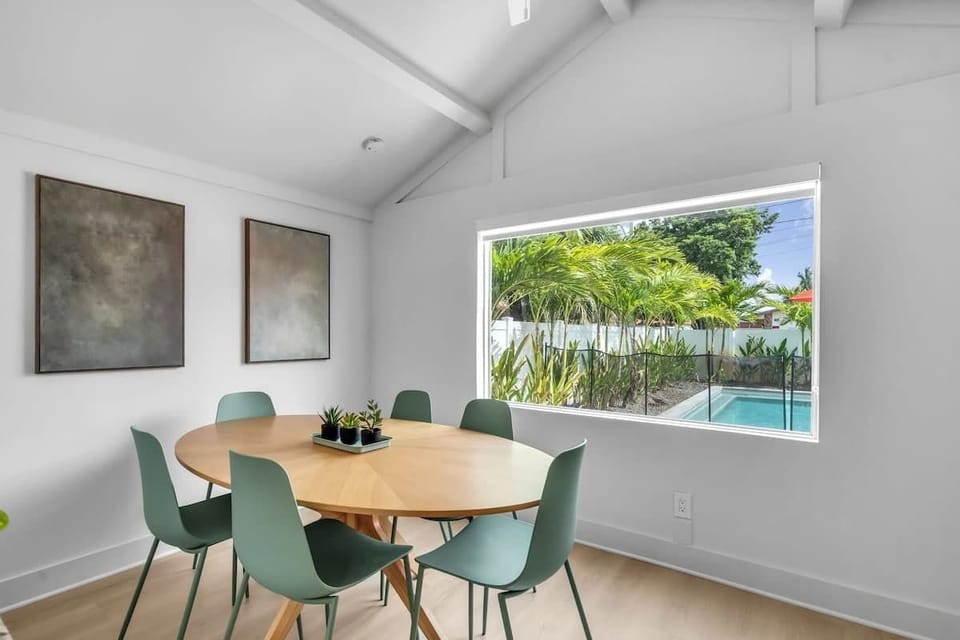 Bright dining space with a round table for six, vaulted ceilings, and a large window framing the pool and tropical yard. Ideal for gathering over meals with a view.