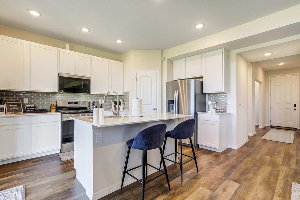 White shaker cabinetry surrounds a quartz topped island with seating for three, styled with black barstools. A stainless microwave and hardwood style flooring complete this crisp, modern kitchen.